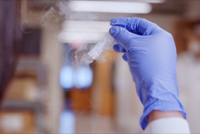 A close-up image of a biomedical researcher wearing rubber gloves and holding a vial containing samples